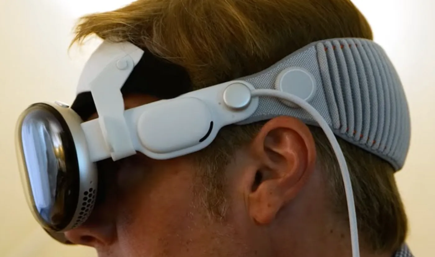 Profile view of a man wearing the Apple Vision Pro with the Cognixion Open Face Comfort System: a white bracket and knit headband holding an EEG sensor above and behind the ear, with a thin cable running down the neck.